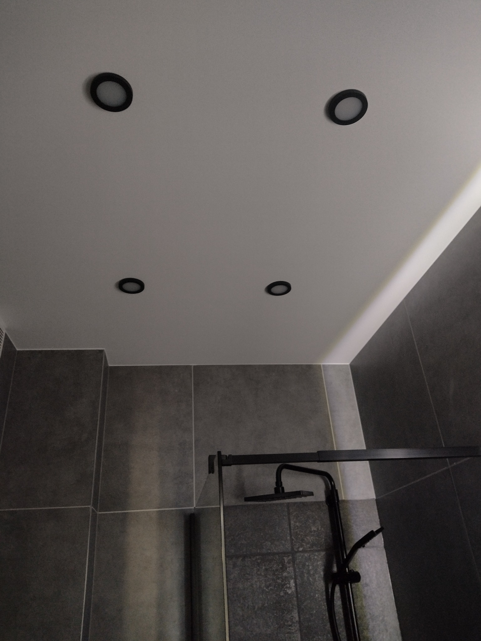 Nowoczesna łazienka z szarymi płytkami, czarną armaturą i oświetleniem punktowym. Minimalistyczny design, kabina prysznicowa z czarnymi profilami.