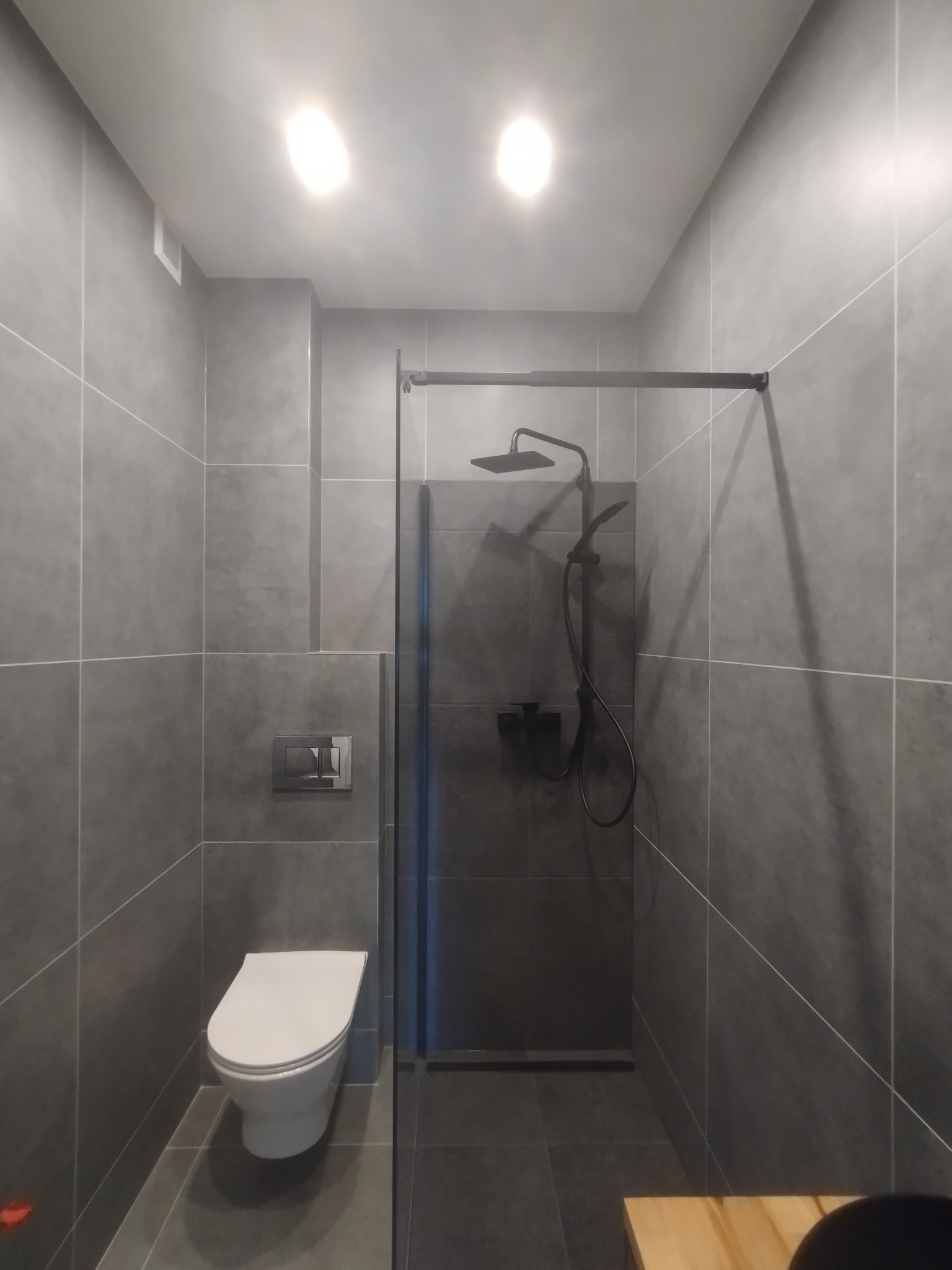 Nowoczesna łazienka w szarościach z kabiną typu walk-in i czarną armaturą. Uwagę zwraca minimalistyczna toaleta podwieszana i duże, kwadratowe płytki.