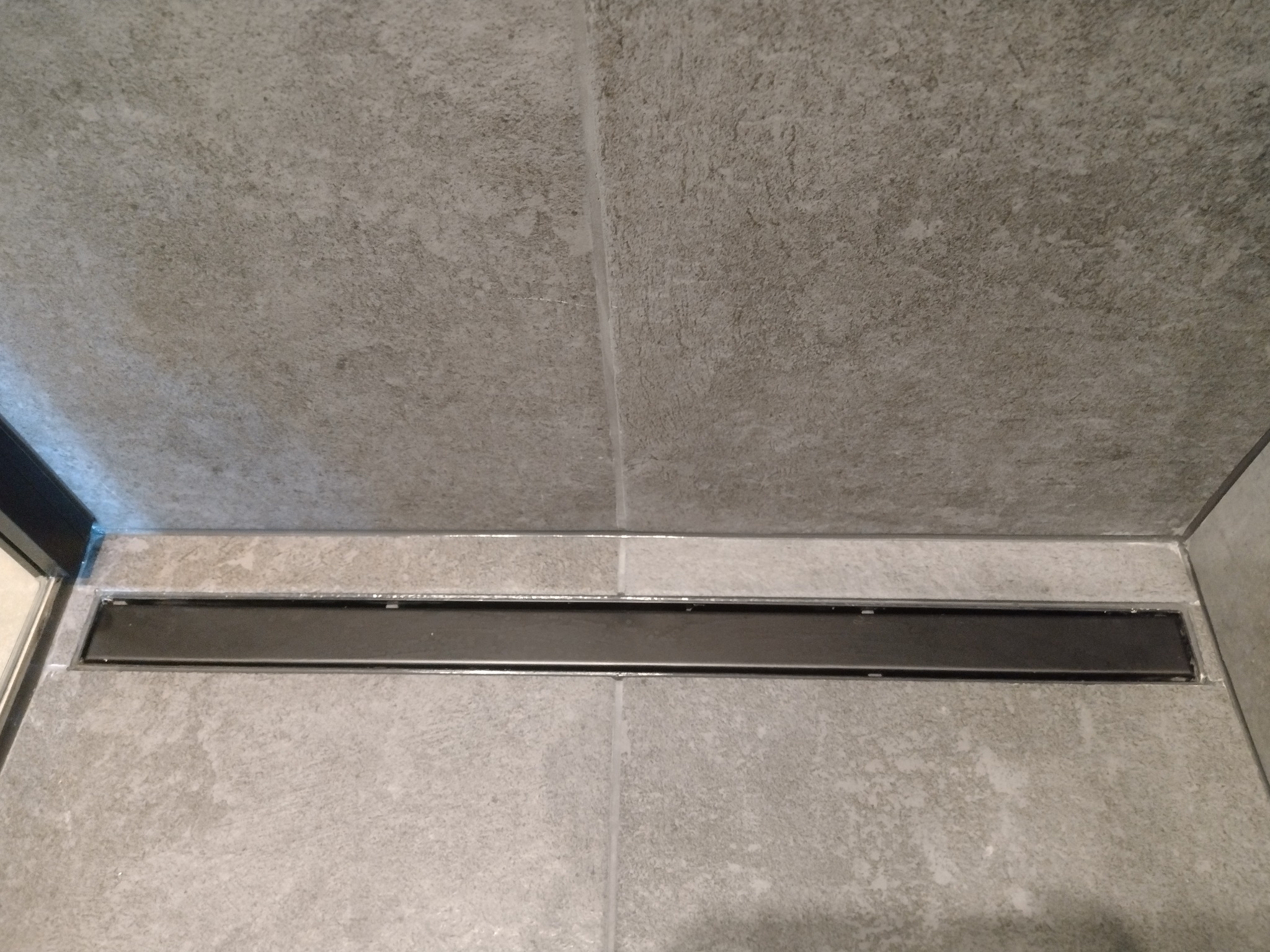 Szczegółowe ujęcie liniowego odpływu prysznicowego wkomponowanego w szare, matowe płytki ceramiczne. Minimalistyczny design, precyzyjne wykończenie, nowoczesna łazienka.