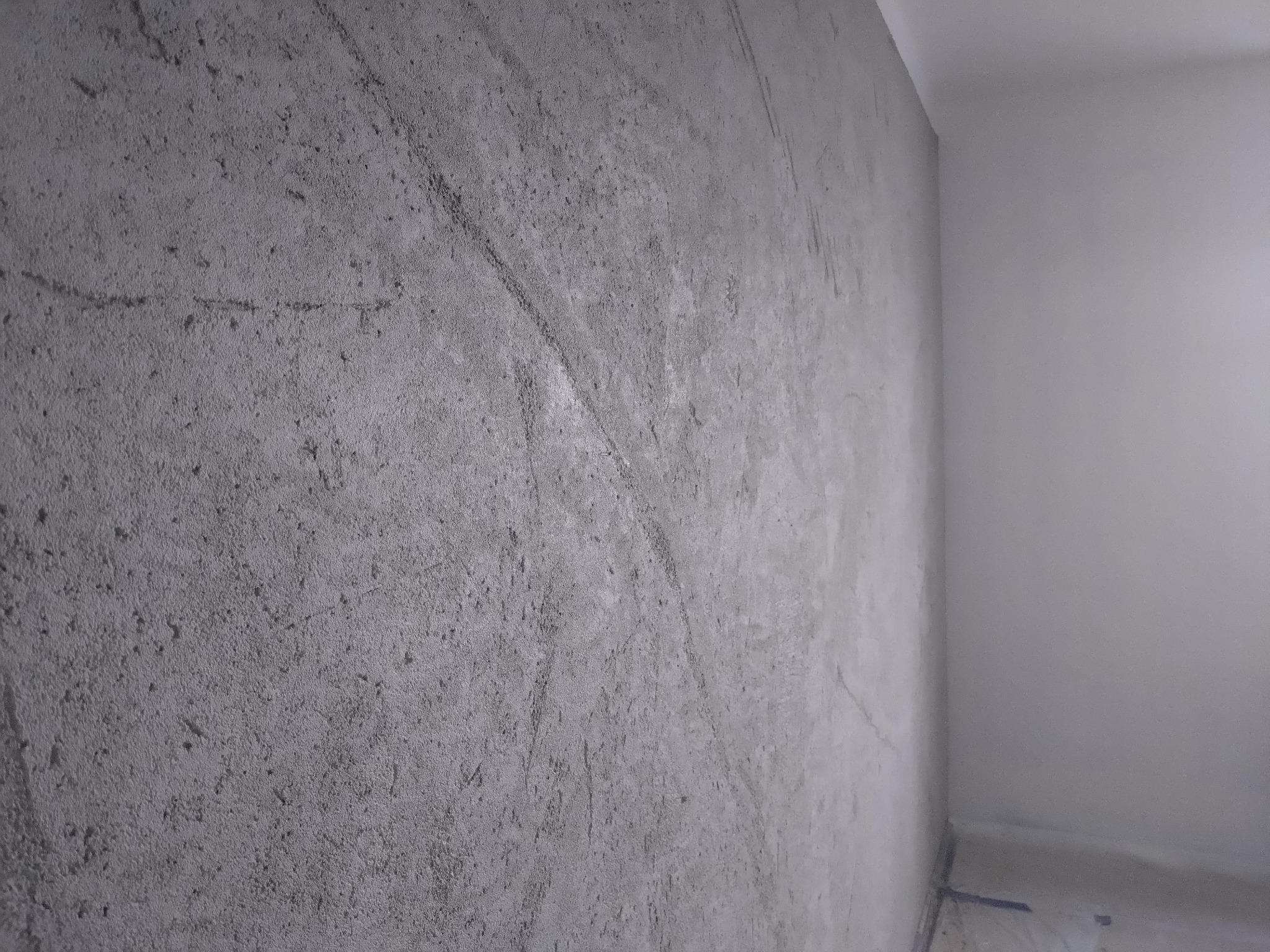 Ściana z szarego tynku dekoracyjnego imitującego beton, z widoczną fakturą i delikatnymi smugami, w rogu pomieszczenia z białą ścianą. Ujęcie z bliska.