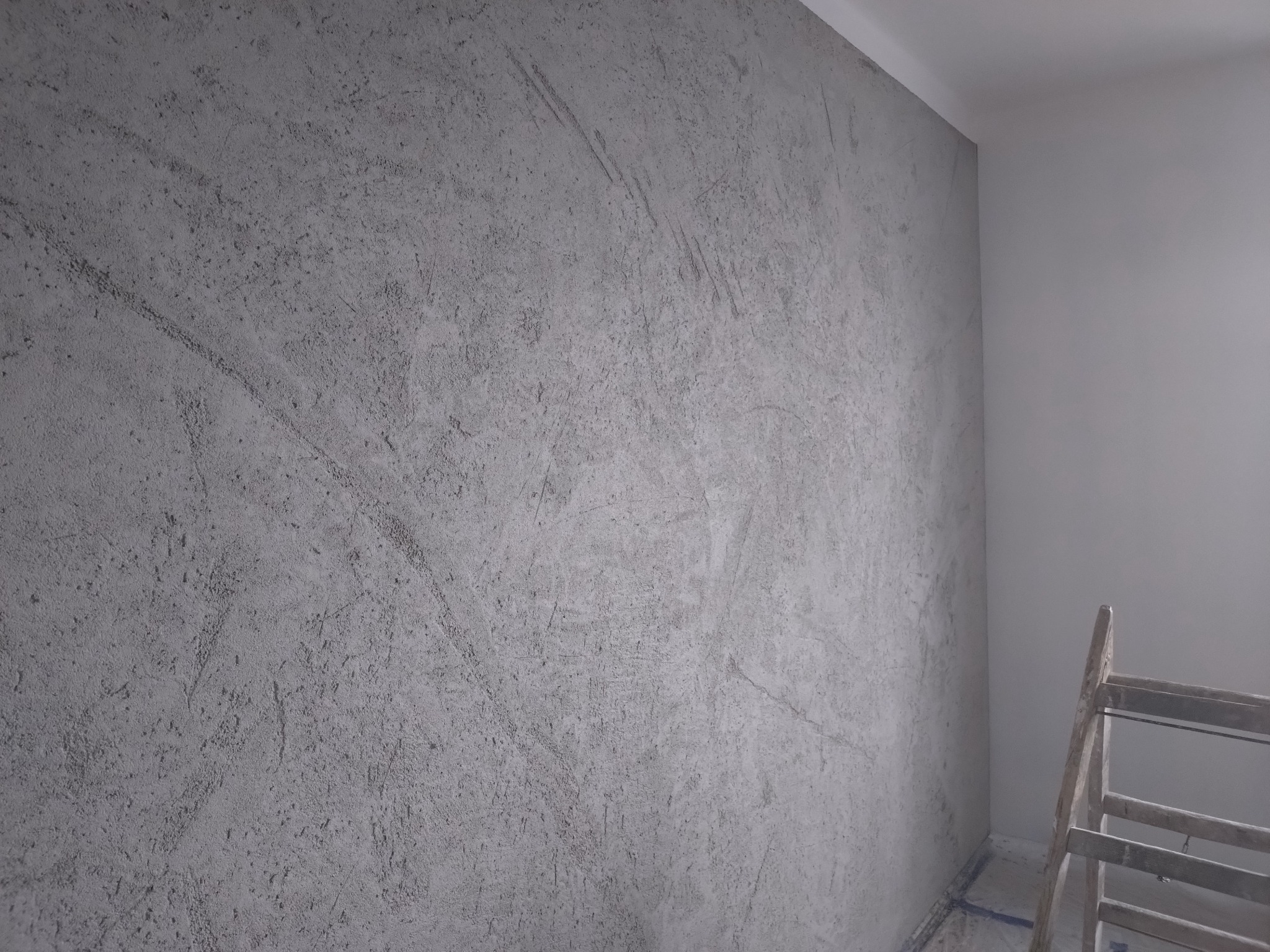 Ściana z szarą, dekoracyjną fakturą betonu, widoczna z bliska, z fragmentem drabiny malarskiej w rogu. Tynk dekoracyjny w neutralnej tonacji.