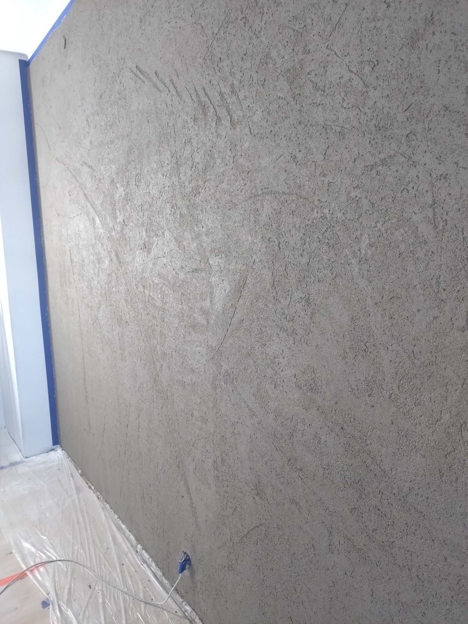 Ściana z szarym tynkiem dekoracyjnym imitującym beton, zabezpieczona folią malarską przy podłodze. Widoczne ślady po narzędziach i charakterystyczna faktura betonu.