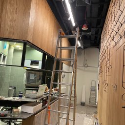 extremePROJECT Alicja Świerczyńska - Montaż drewnianej okładziny ściennej w lokalu użytkowym, widoczna drabina, narzędzia i materiały budowlane, surowy stan wykończenia.