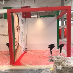 extremePROJECT Alicja Świerczyńska - Stoisko targowe w trakcie montażu, czerwono-zielona konstrukcja, plakaty z wizerunkiem kobiety, czarne hokery, podłoga zabezpieczona folią, paleta z towarem w folii.