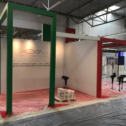 extremePROJECT Alicja Świerczyńska - Stoisko targowe w trakcie montażu, z zielonymi i czerwonymi elementami konstrukcyjnymi, podłoga zabezpieczona folią, widoczne krzesła barowe i paleta z materiałami.