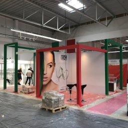 extremePROJECT Alicja Świerczyńska - Przygotowania do targów w Warszawie: montaż stoiska z czerwono-zieloną konstrukcją, na podłodze folia ochronna, w tle widoczni pracownicy.