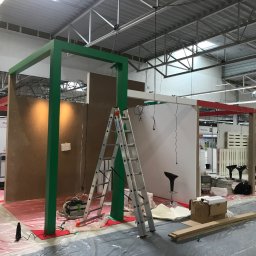 extremePROJECT Alicja Świerczyńska - Montaż stoiska targowego: widoczne elementy konstrukcyjne pomalowane na zielono i czerwono, płyty meblowe, drabina aluminiowa, folia ochronna na podłodze, narzędzia i materiały budowlane, hala...