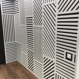 extremePROJECT Alicja Świerczyńska - Geometryczna dekoracja ścienna z białych listewek na czarnym tle, tworząca wzory pionowe, poziome i ukośne, widoczny fragment drewnianej podłogi i włącznik światła.