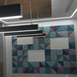 Akustyczna ściana z trójkątnych paneli w odcieniach niebieskiego, różu i czerwieni, z białymi przestrzeniami, oświetlona przez nowoczesne, podłużne lampy sufitowe.