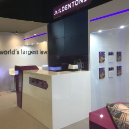 extremePROJECT Alicja Świerczyńska - Nowoczesne stoisko targowe z białym biurkiem recepcyjnym, logiem firmy Dentons, oświetleniem LED w kolorze fioletowym i ścianą z plakatami informacyjnymi.