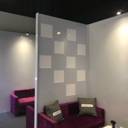 extremePROJECT Alicja Świerczyńska - Fragment aranżacji wnętrza z fioletową sofą, dekoracyjnymi poduszkami z napisem i ścianą z kwadratowymi wnękami, oświetlonej punktowo.