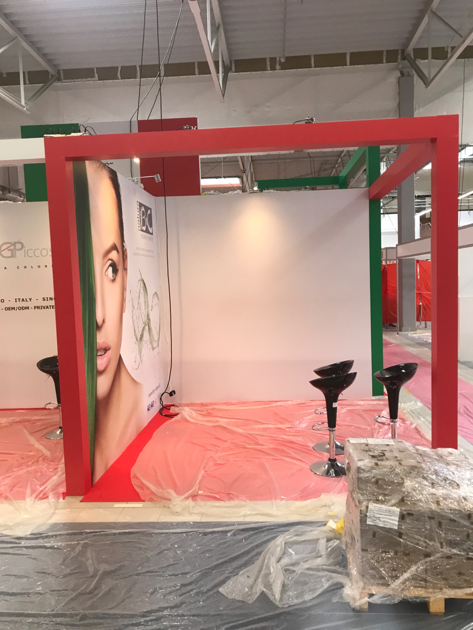 Stoisko targowe w trakcie montażu, czerwono-zielona konstrukcja, plakaty z wizerunkiem kobiety, czarne hokery, podłoga zabezpieczona folią, paleta z towarem w folii.