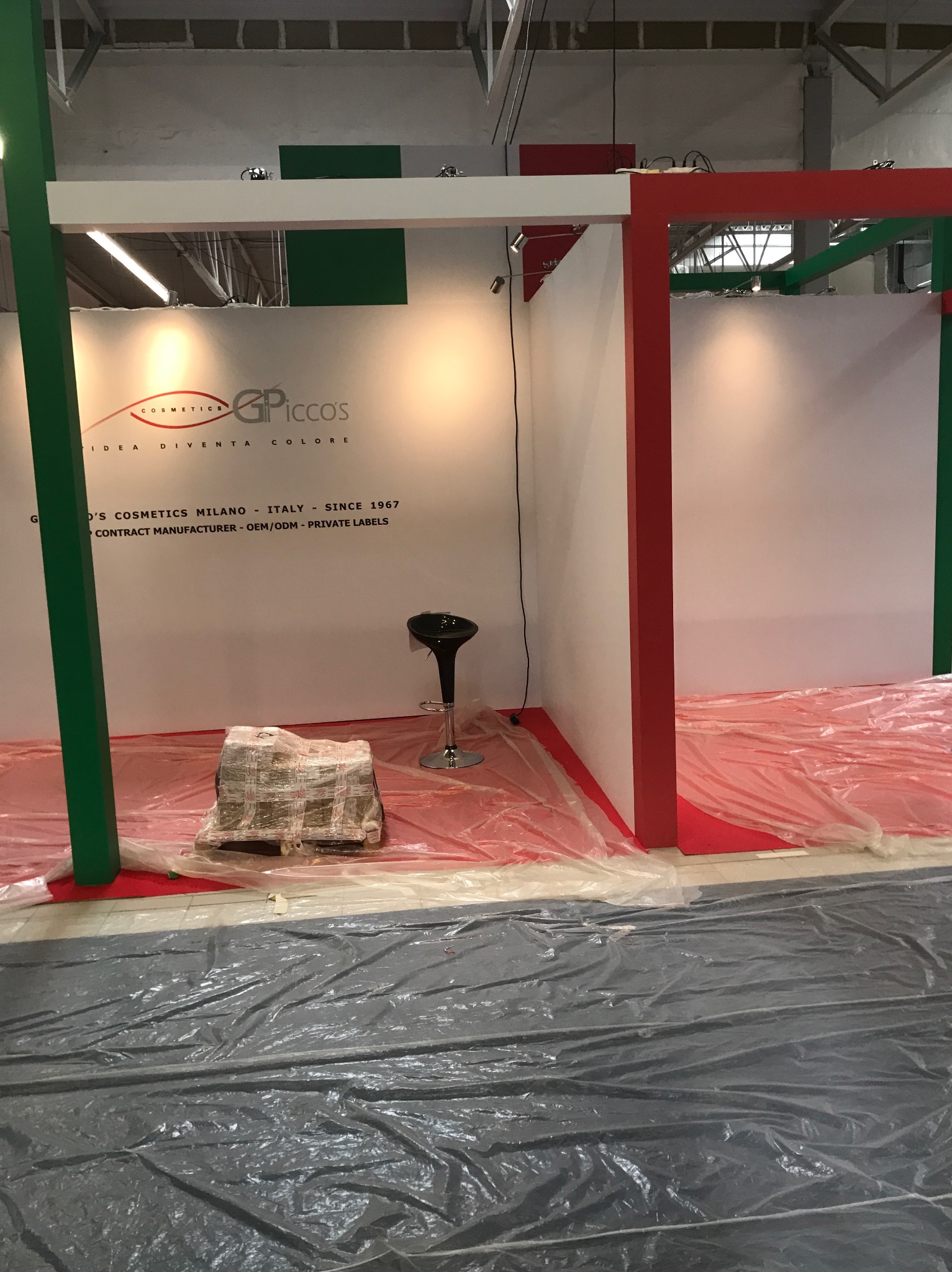 Przygotowania do targów: stoisko kosmetyczne w trakcie montażu, widoczne elementy konstrukcyjne w kolorach czerwonym i zielonym, zabezpieczona folią podłoga, krzesło barowe i zapakowany produkt...