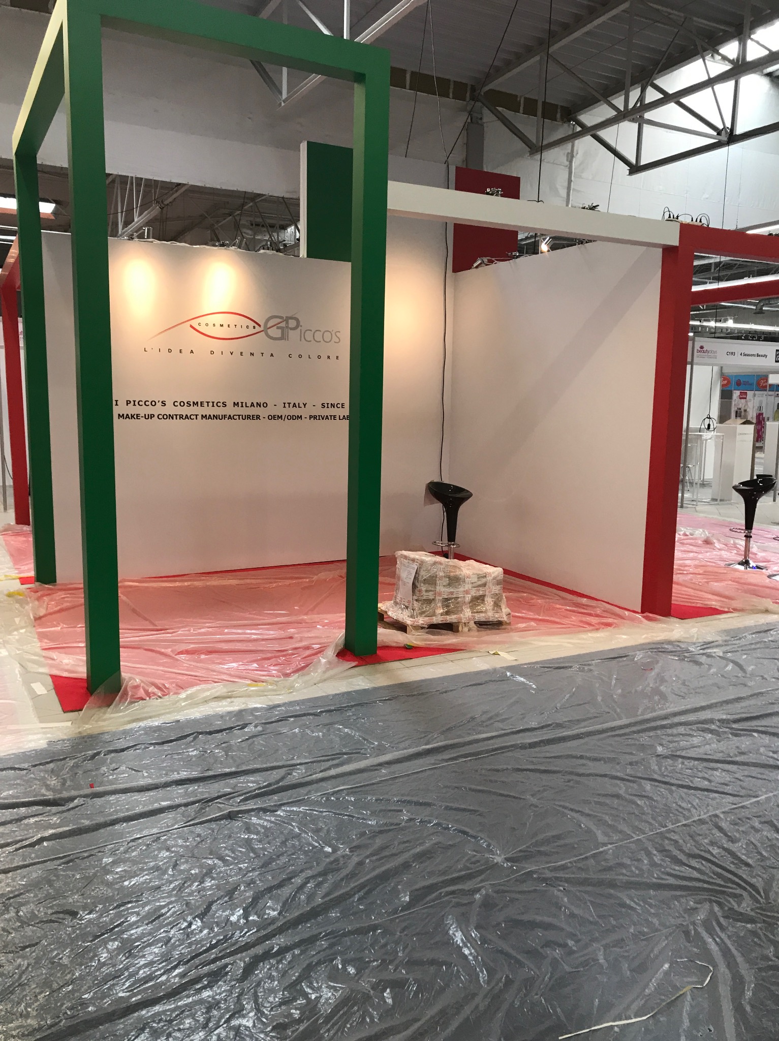 Stoisko targowe w trakcie przygotowań: widoczne elementy konstrukcyjne w kolorach zielonym i czerwonym, logo firmy kosmetycznej na białej ścianie, podłoga zabezpieczona folią ochronną, dwa czarne...