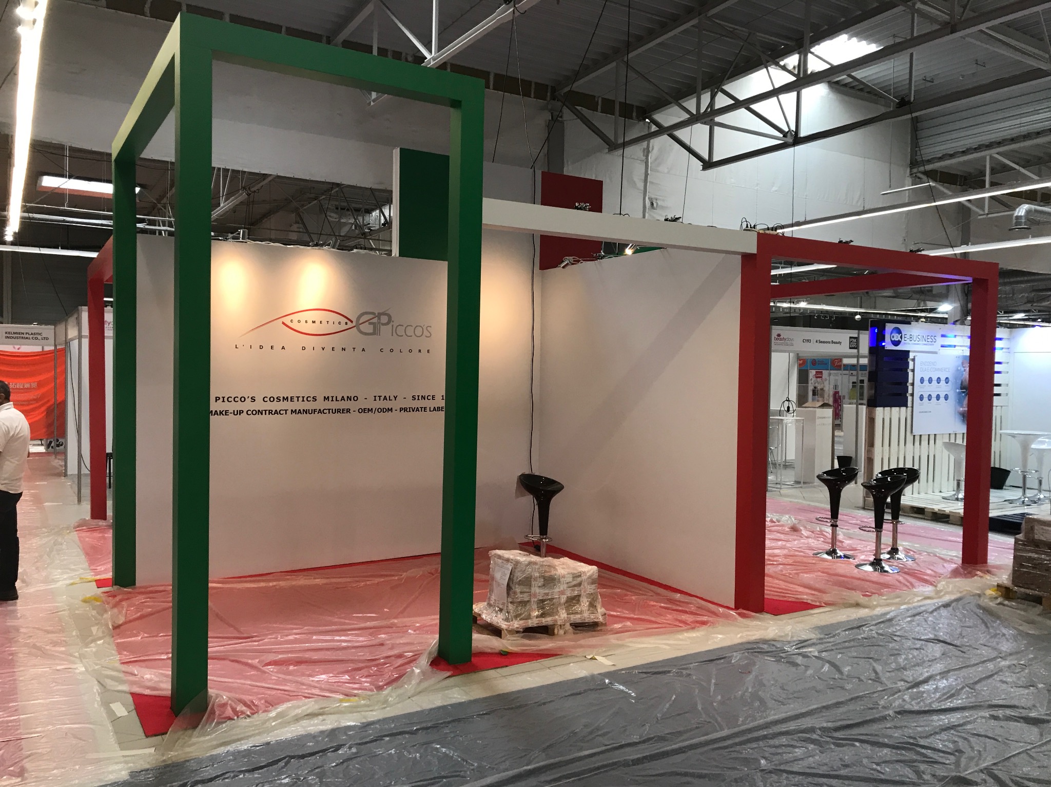 Stoisko targowe w trakcie montażu, z zielonymi i czerwonymi elementami konstrukcyjnymi, podłoga zabezpieczona folią, widoczne krzesła barowe i paleta z materiałami.