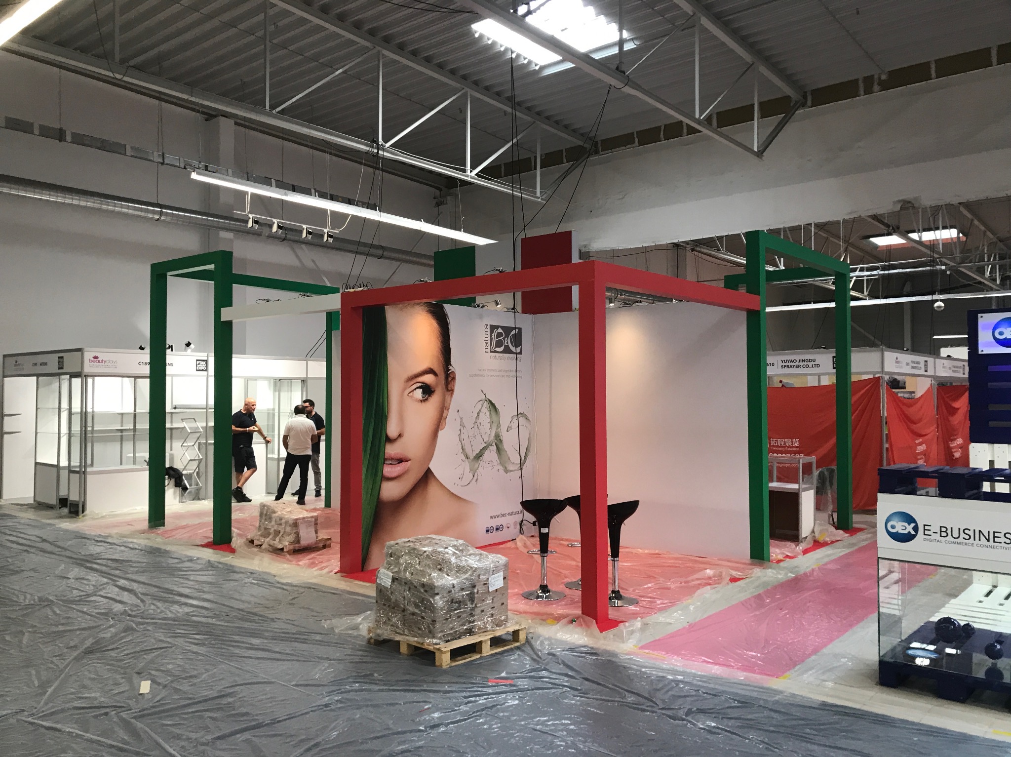 Przygotowania do targów w Warszawie: montaż stoiska z czerwono-zieloną konstrukcją, na podłodze folia ochronna, w tle widoczni pracownicy.