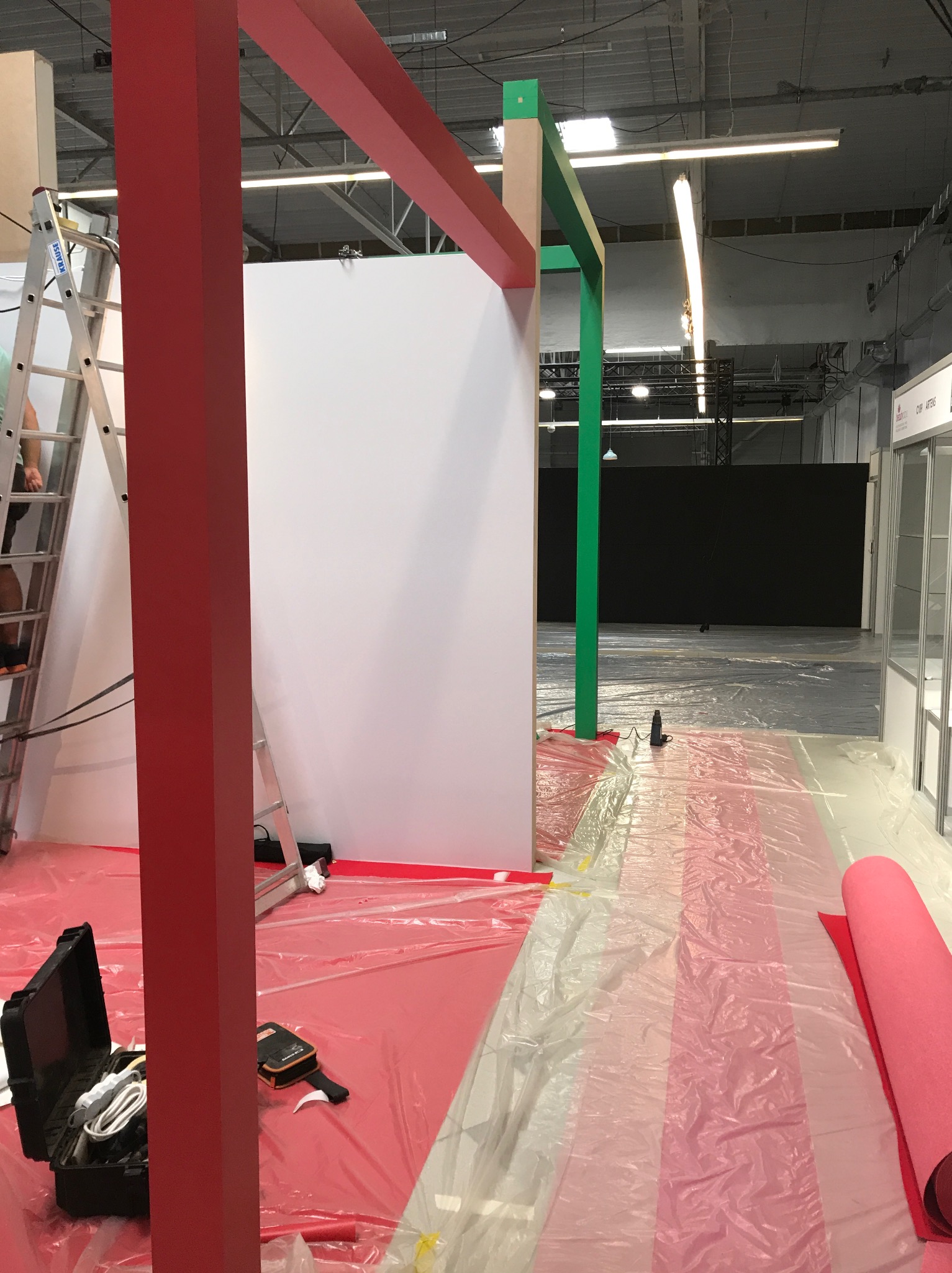 Montaż stoiska targowego: czerwone i zielone ramy konstrukcyjne, drabina oparta o białą ścianę, narzędzia w walizce, folia ochronna na podłodze.