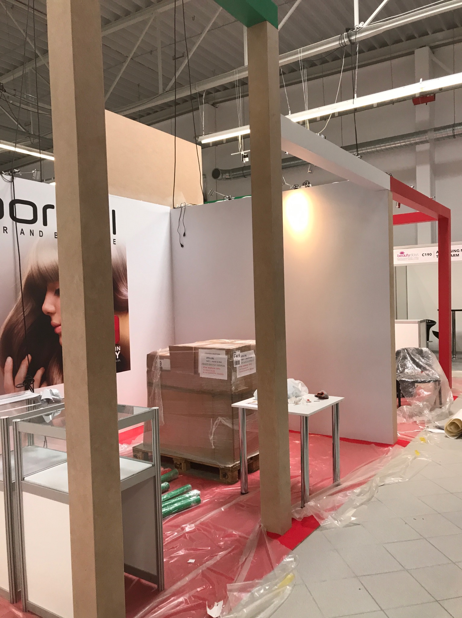 Budowa stoiska targowego w trakcie montażu: widoczne surowe drewniane słupy konstrukcyjne, fragment ekspozycji z grafiką kobiety, zapakowane kartony na palecie, małe stoliki i podłoga zabezpieczona...