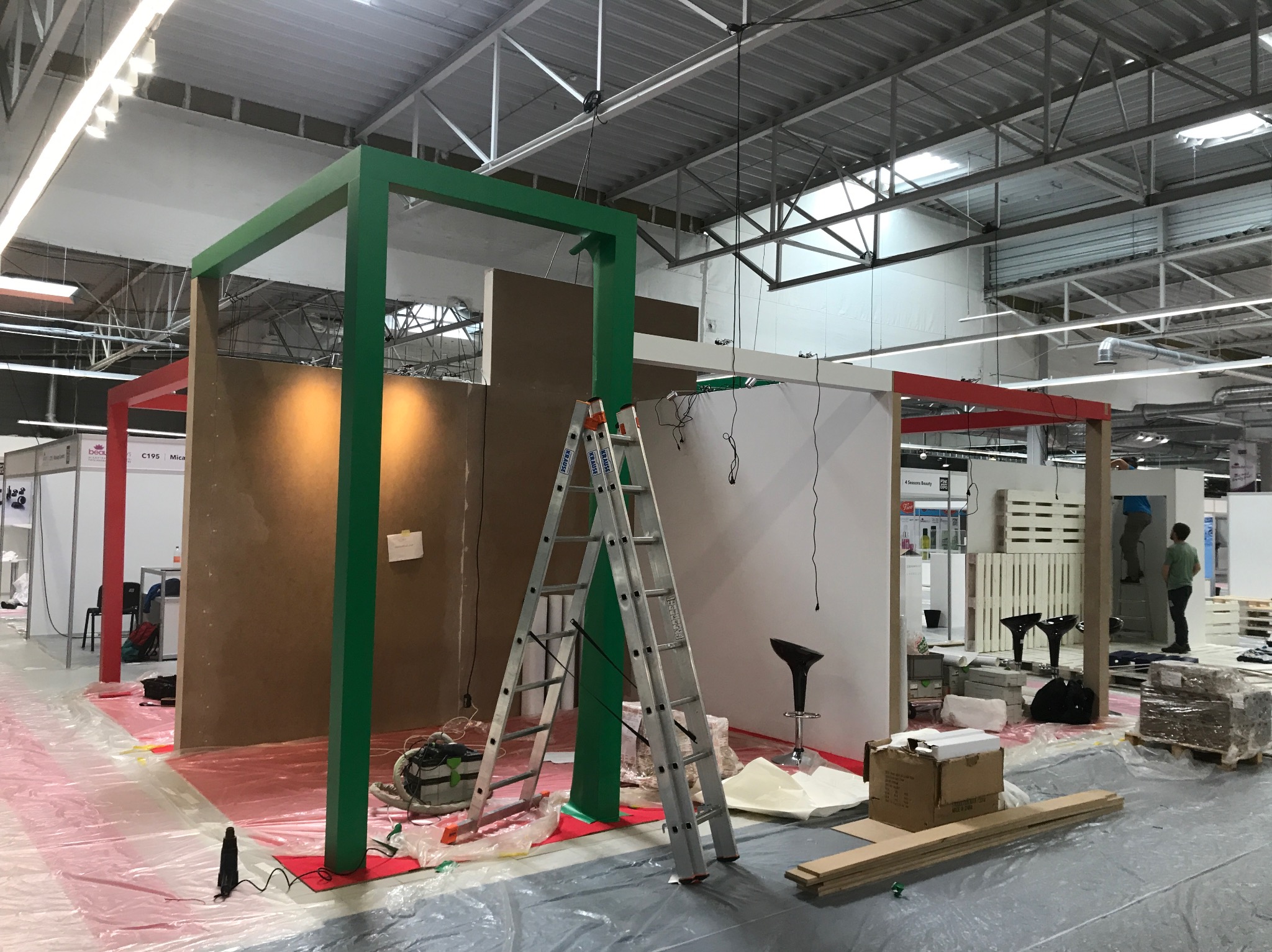 Montaż stoiska targowego: widoczne elementy konstrukcyjne pomalowane na zielono i czerwono, płyty meblowe, drabina aluminiowa, folia ochronna na podłodze, narzędzia i materiały budowlane, hala...