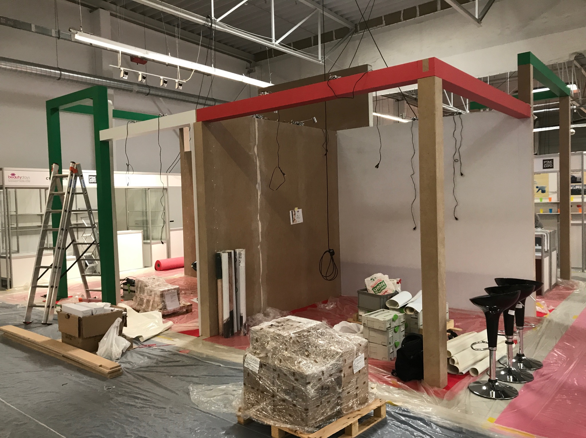 Montaż stoiska targowego w hali: drewniane i metalowe konstrukcje w kolorach czerwonym, zielonym i białym, zabezpieczone folią podłogi, palety z materiałami i narzędzia w nieładzie.