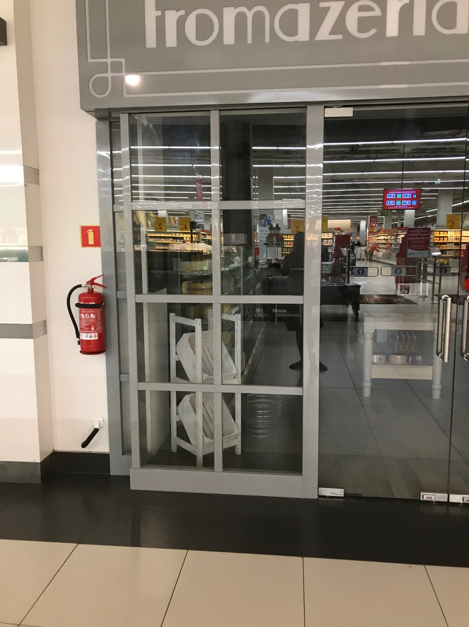 Wejście do sklepu 'fromazeria' z przesuwnymi szklanymi drzwiami i widocznym wnętrzem sklepu z regałami i klientami, na ścianie gaśnica.