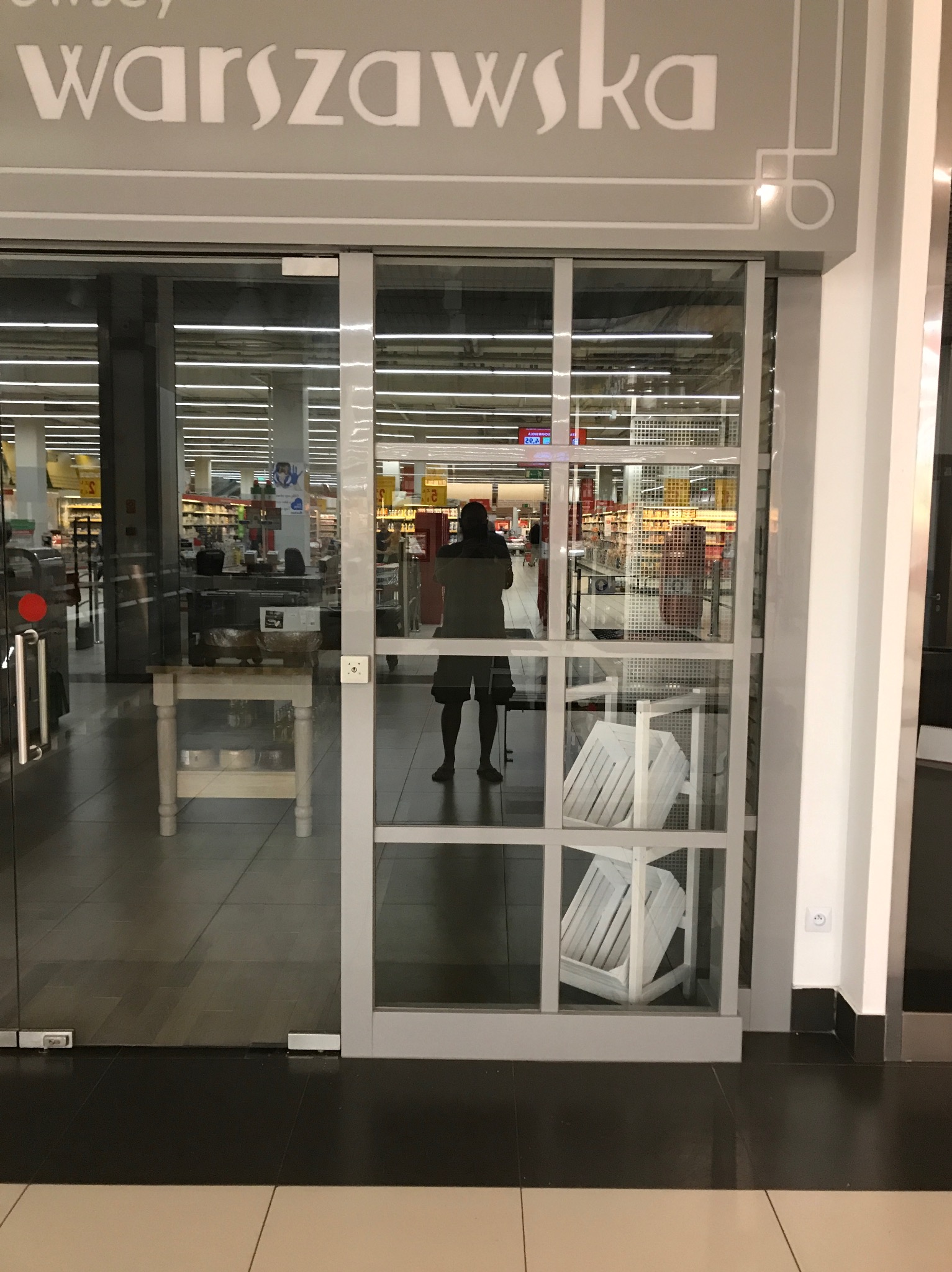 Wejście do sklepu 'Warszawska' z przesuwanymi drzwiami szklanymi w metalowej ramie, w tle widoczne półki sklepowe i osoba stojąca tyłem, wewnątrz sklepu stolik z ekspozycją produktów i białe...