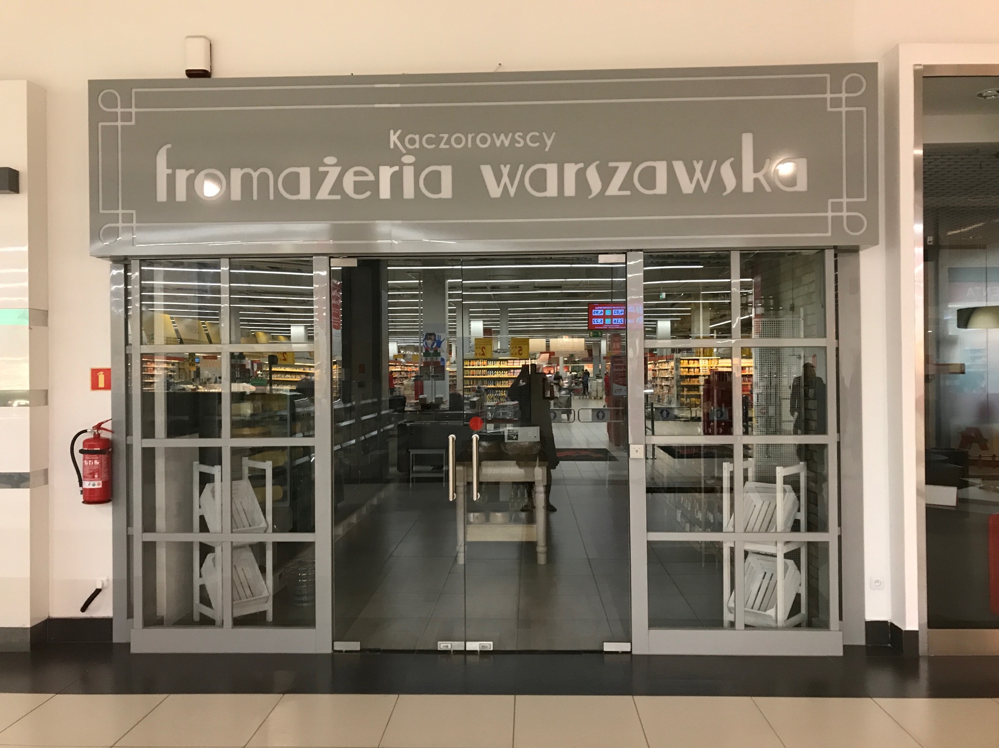Wejście do sklepu 'Fromażeria Warszawska Kaczorowscy' z szklanymi drzwiami, na których widoczne są białe drewniane skrzynki dekoracyjne, a w tle regały z produktami spożywczymi.