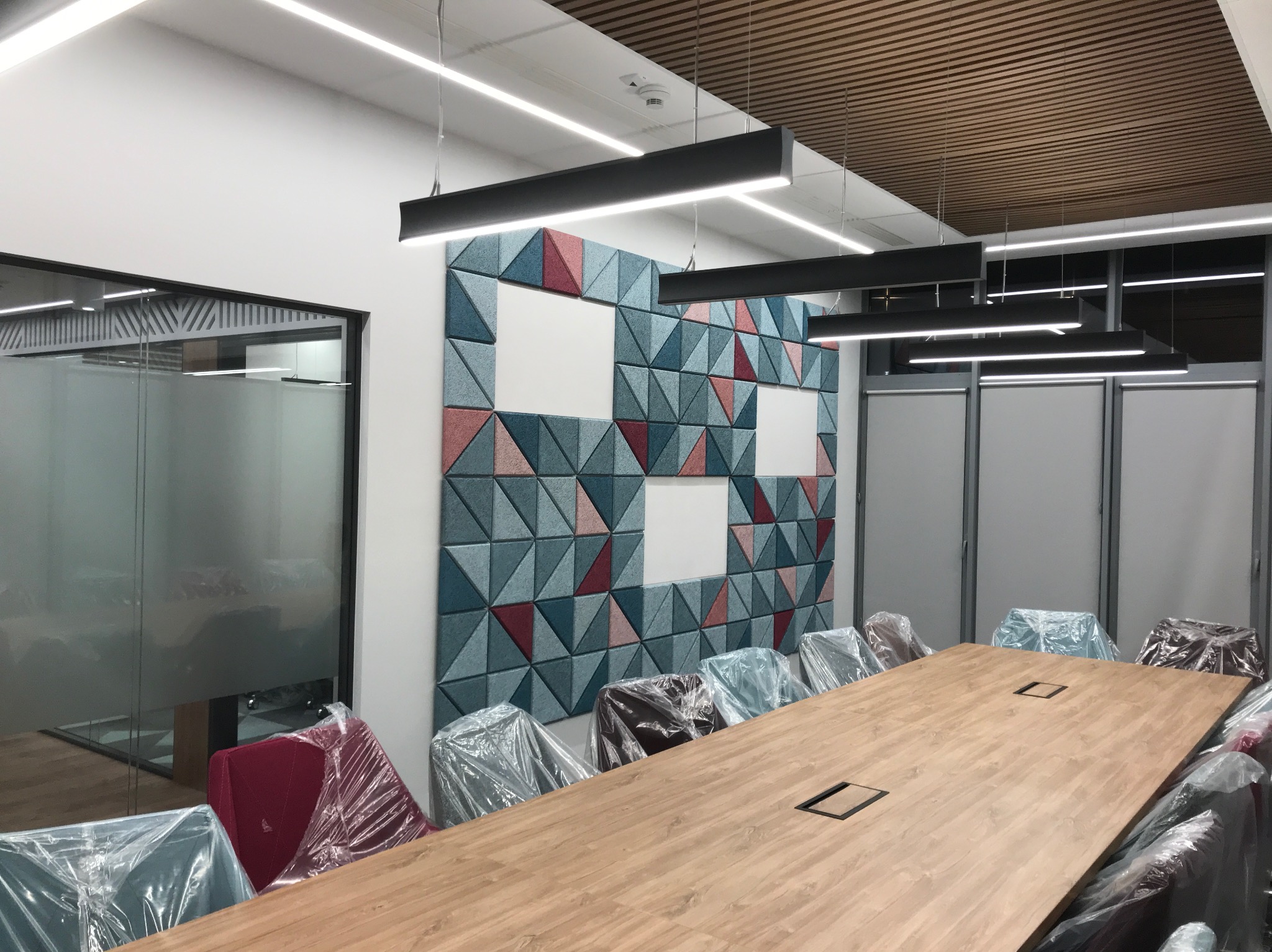 Nowoczesna sala konferencyjna z długim drewnianym stołem, krzesłami w foliowych pokrowcach, oświetleniem LED i dekoracyjnym panelem ściennym z trójkątów w odcieniach niebieskiego i czerwonego.