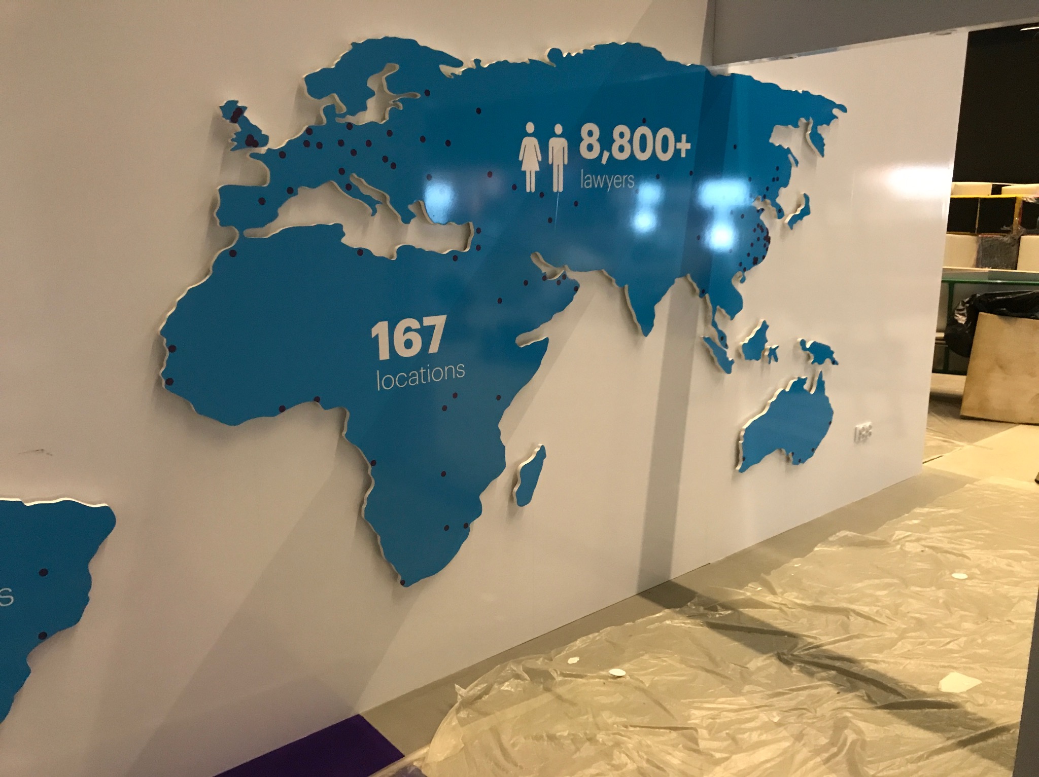 Niebieska mapa świata z zaznaczonymi lokalizacjami i informacją o liczbie prawników (8800+), przymocowana do białej ściany, folia ochronna na podłodze.
