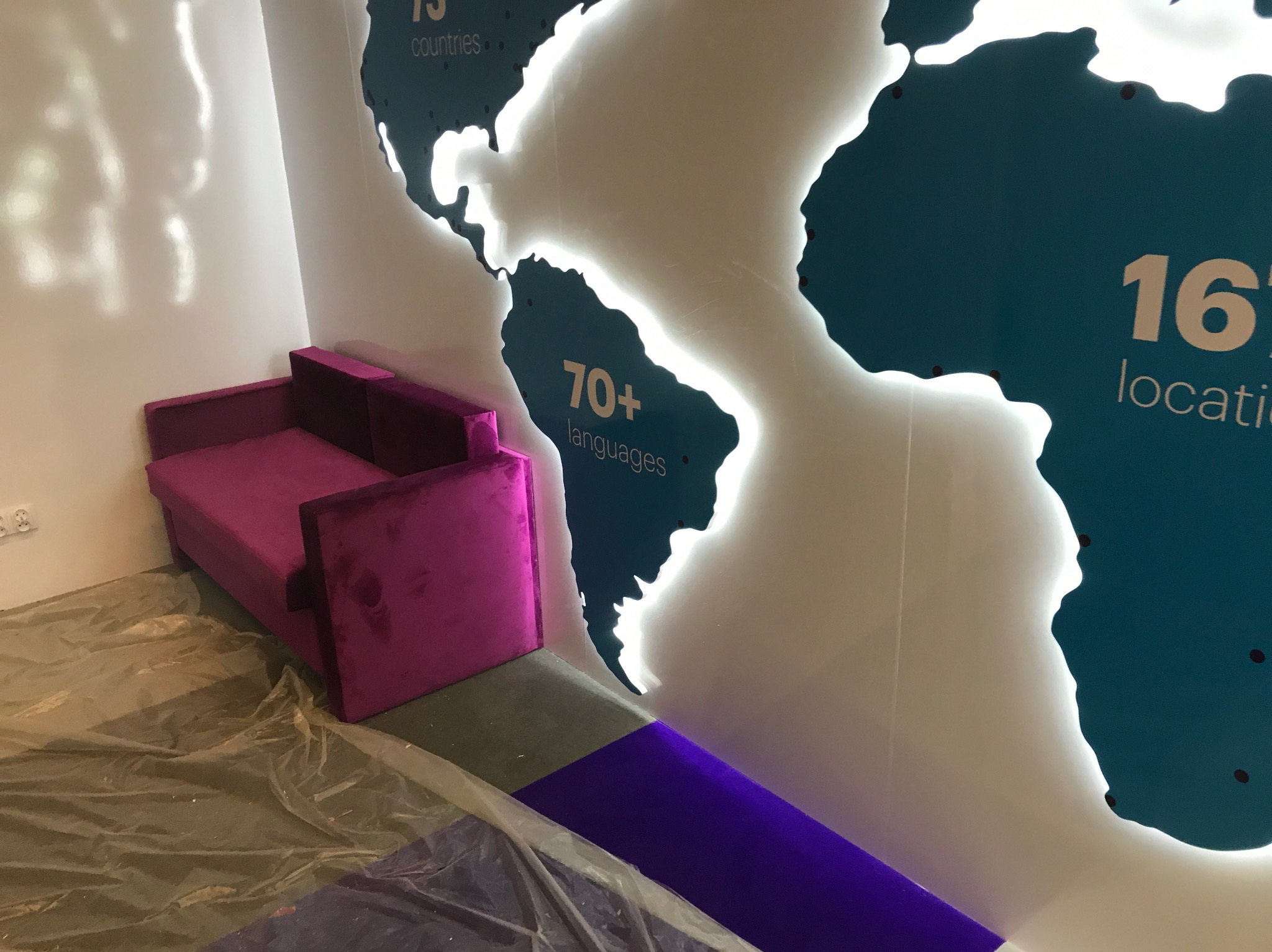 Fioletowa sofa z aksamitną tapicerką stoi przy ścianie z podświetlaną mapą świata. Na podłodze folia ochronna.