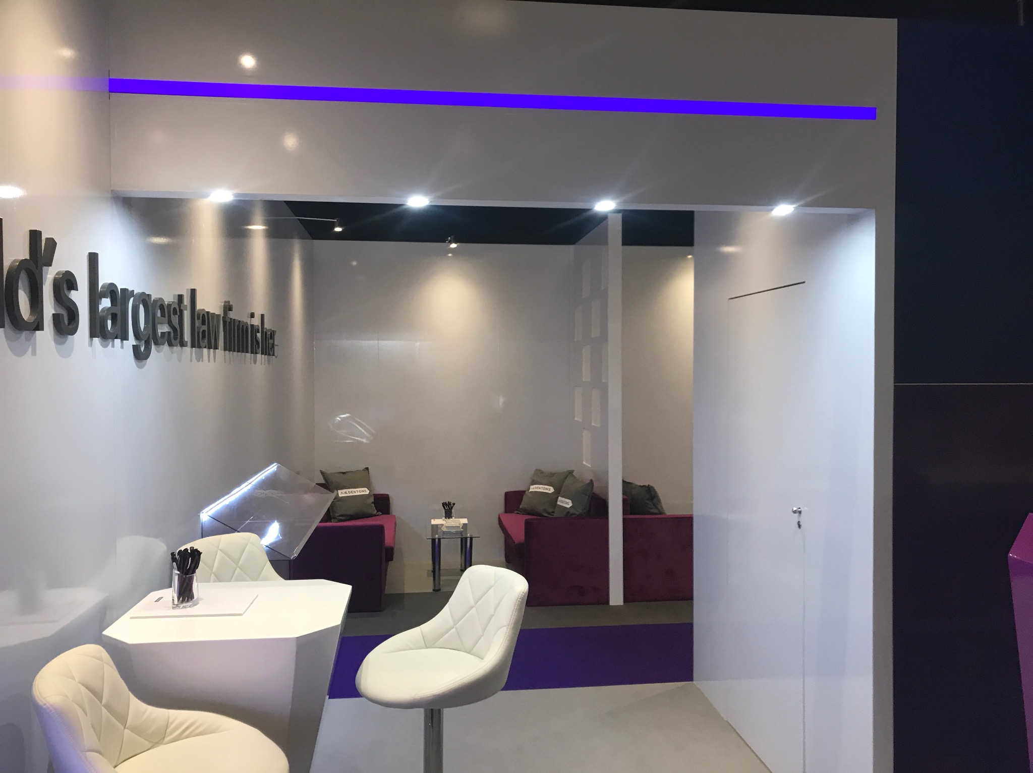 Nowoczesne wnętrze biurowe z białymi ścianami, fioletowym akcentem świetlnym, białym stołem z krzesłami i fioletowymi sofami w tle, oświetlone punktowymi lampami.