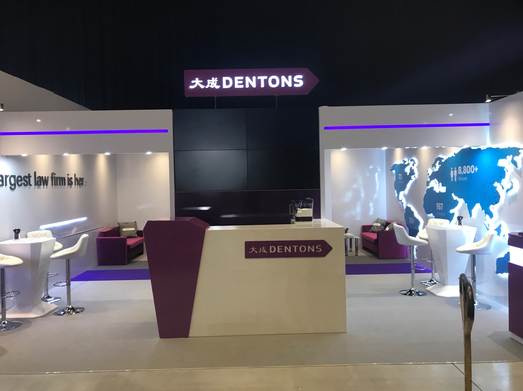 Stoisko targowe firmy Dentons z logo i mapą świata, białe meble barowe i fioletowe akcenty.