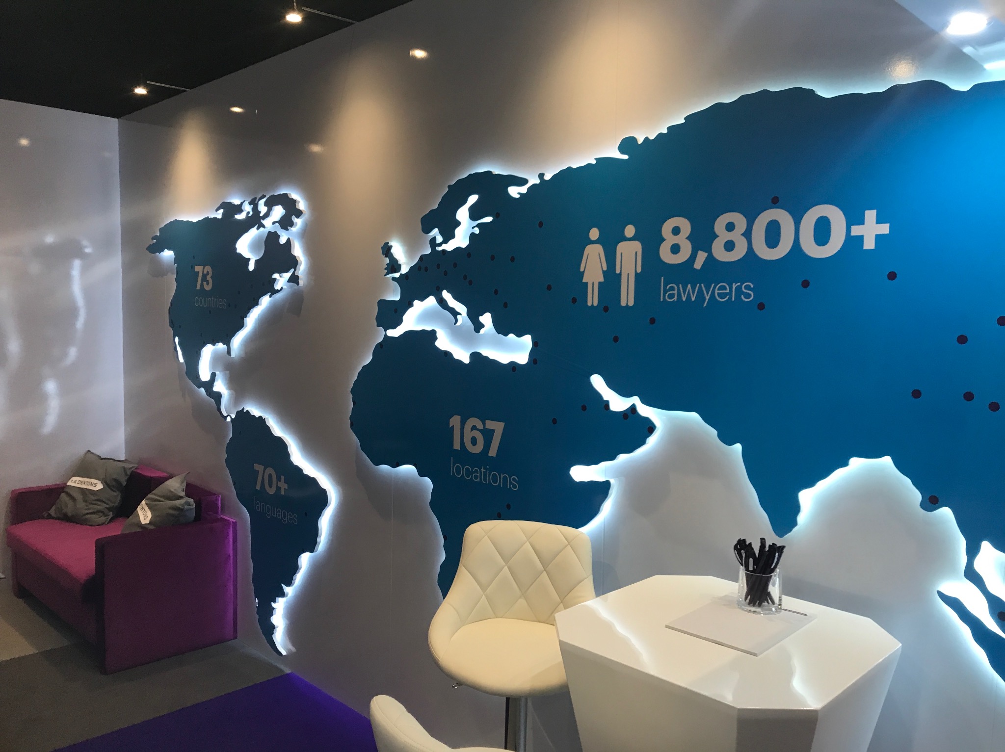 Wnętrze biura z podświetlaną mapą świata na ścianie, informującą o zasięgu firmy: 8,800+ prawników, 167 lokalizacji, 73 kraje, 70+ języków. W tle fioletowa sofa z poduszkami i białe krzesła przy...