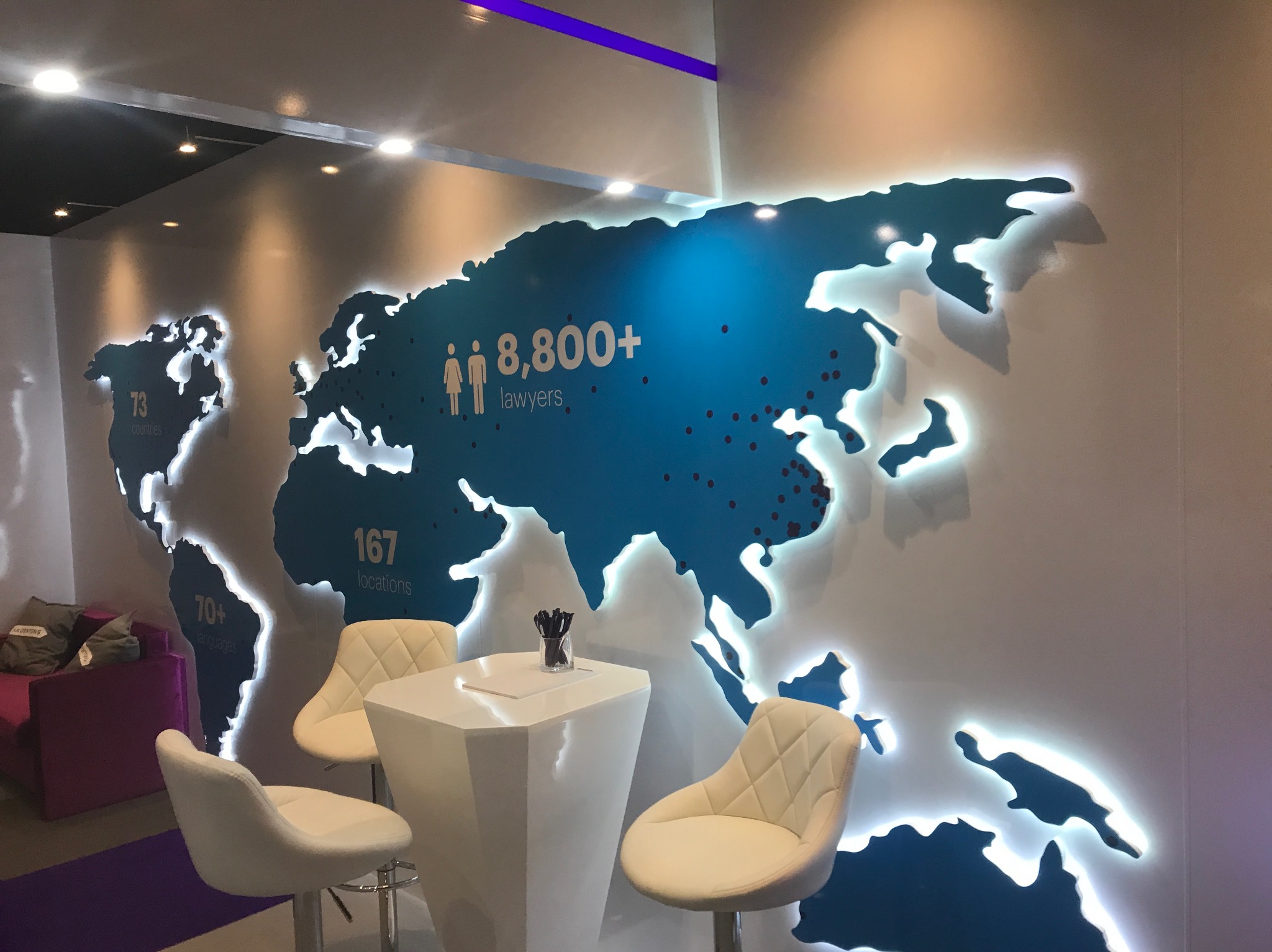 Nowoczesna aranżacja biura z podświetlaną mapą świata, białym stolikiem i krzesłami w stylu skandynawskim, fioletowa sofa w tle.