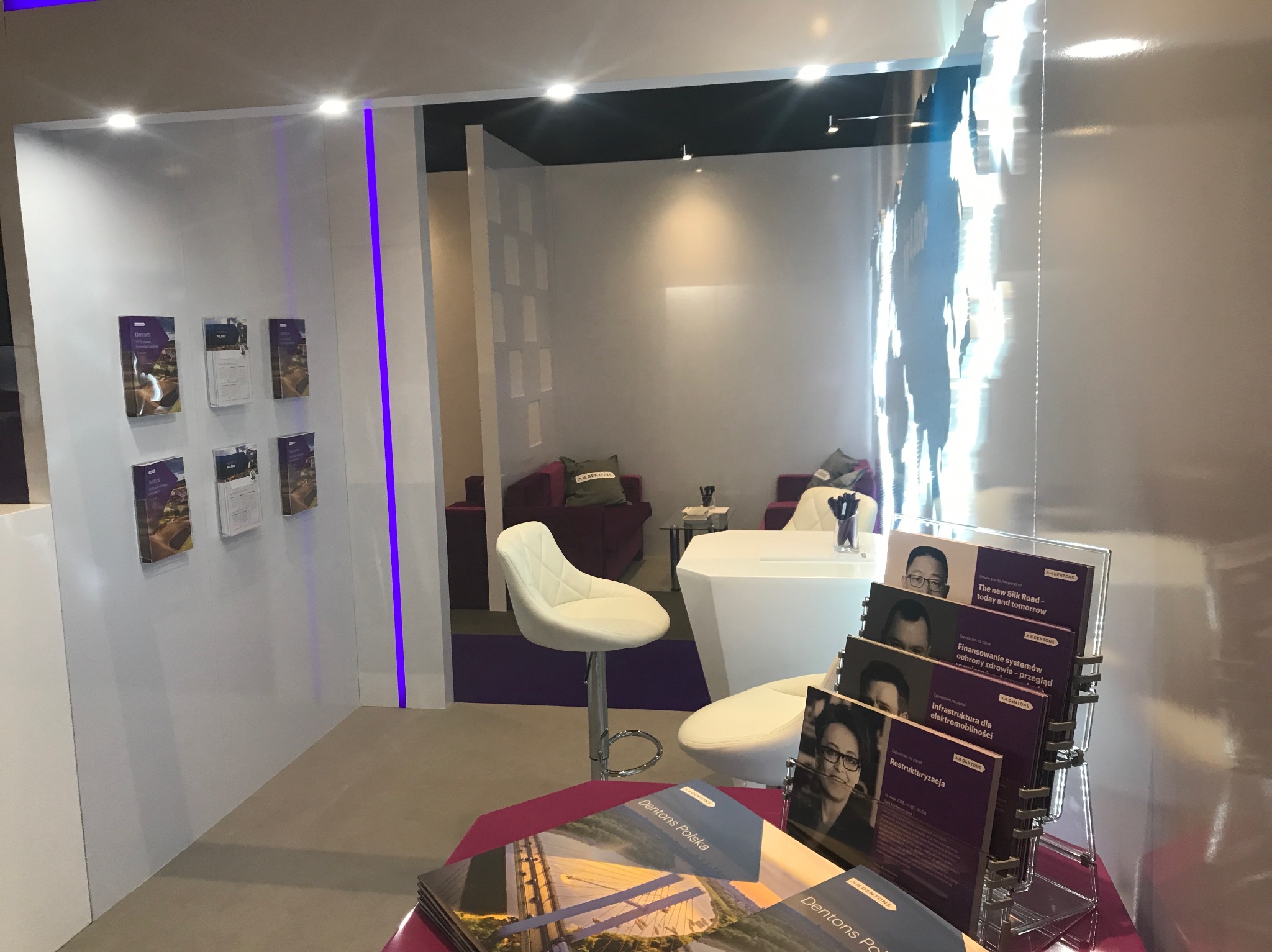 Nowoczesne stoisko targowe z białymi meblami, fioletowymi akcentami i materiałami informacyjnymi Dentons Polska.