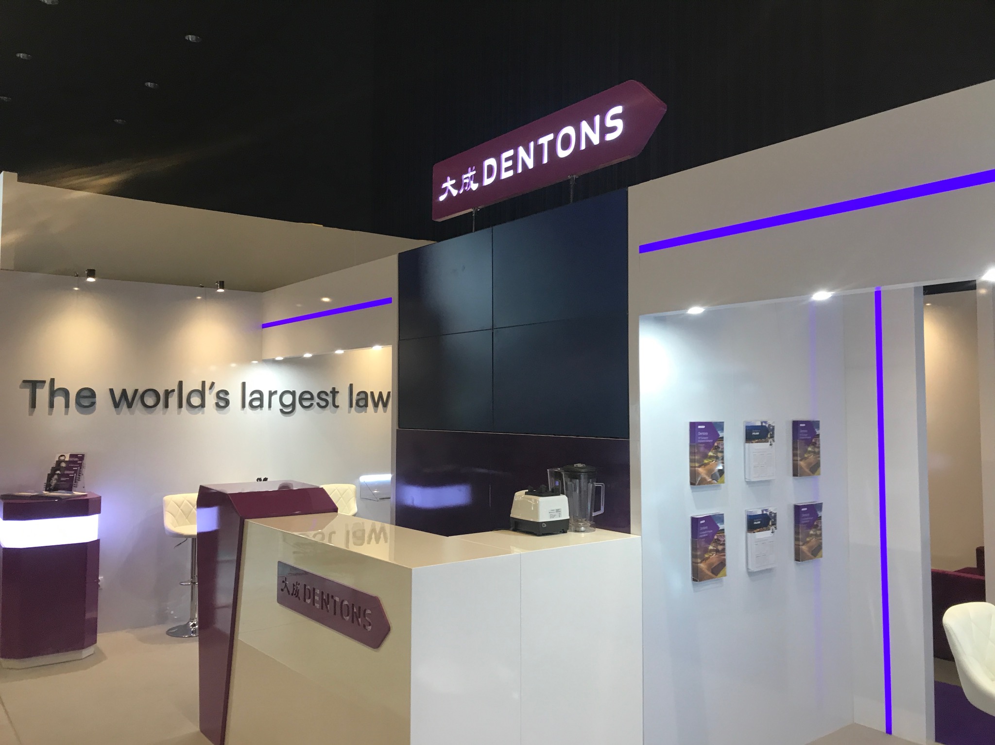 Stoisko targowe firmy Dentons z podświetlanym logo na strzałce, minimalistycznym designem, białymi meblami i fioletowymi akcentami świetlnymi.