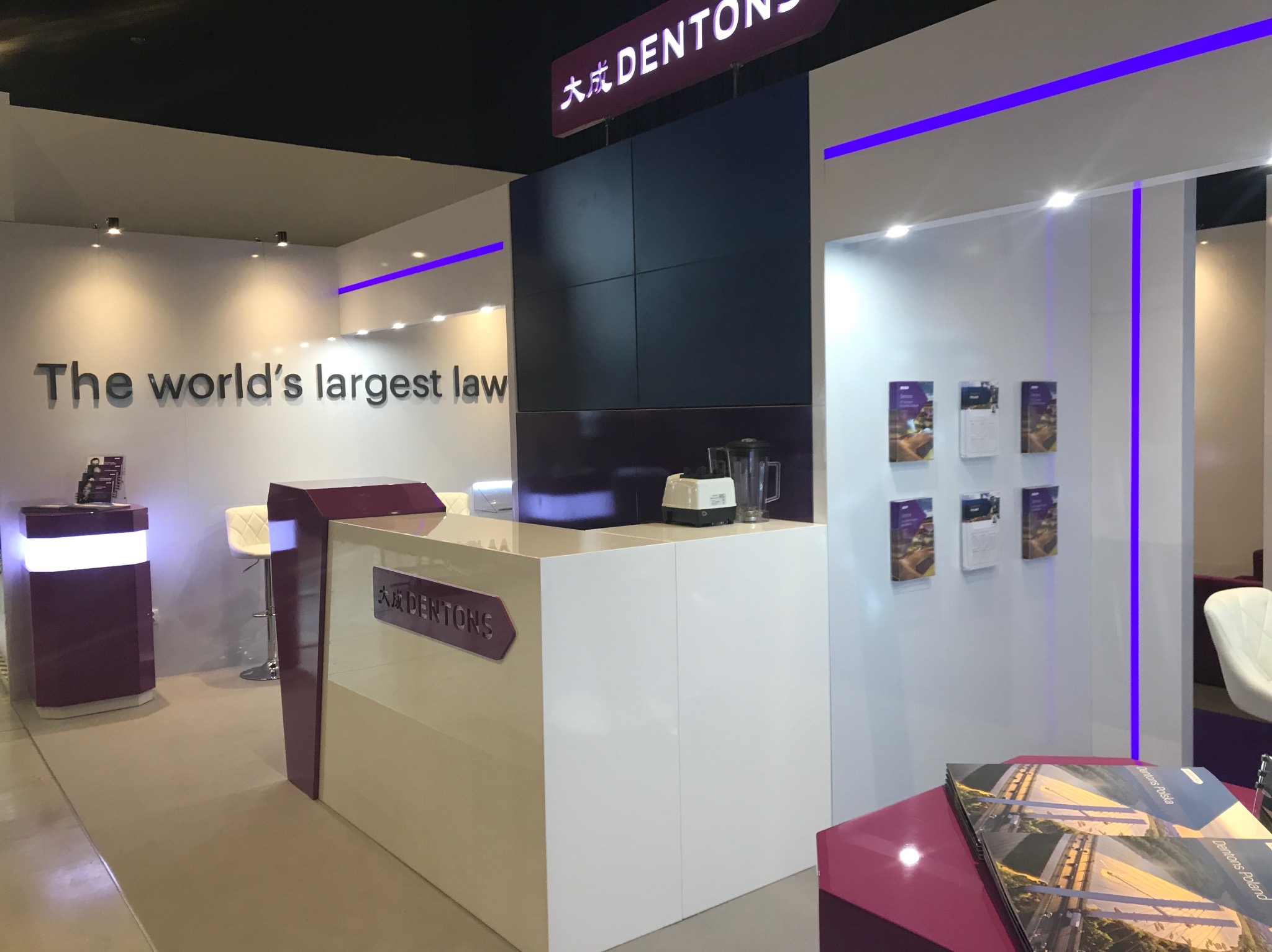 Nowoczesne stoisko targowe z białym biurkiem recepcyjnym, logiem firmy Dentons, oświetleniem LED w kolorze fioletowym i ścianą z plakatami informacyjnymi.