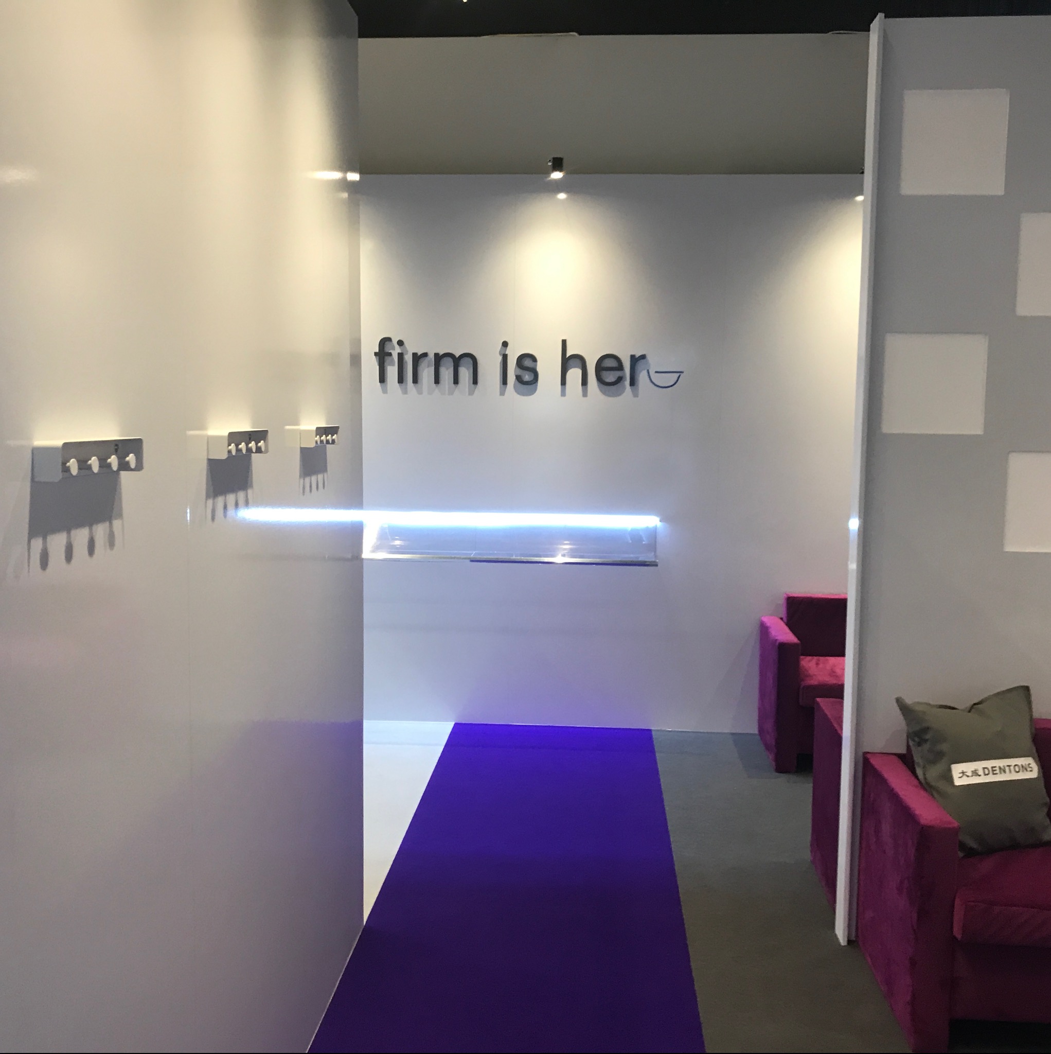 Nowoczesne wnętrze biurowe z białymi ścianami, fioletowym dywanem i elementami dekoracyjnymi, w tym wieszakami na klucze i podświetloną półką, widoczne fotele w kolorze fuksji z poduszką z logo firmy.