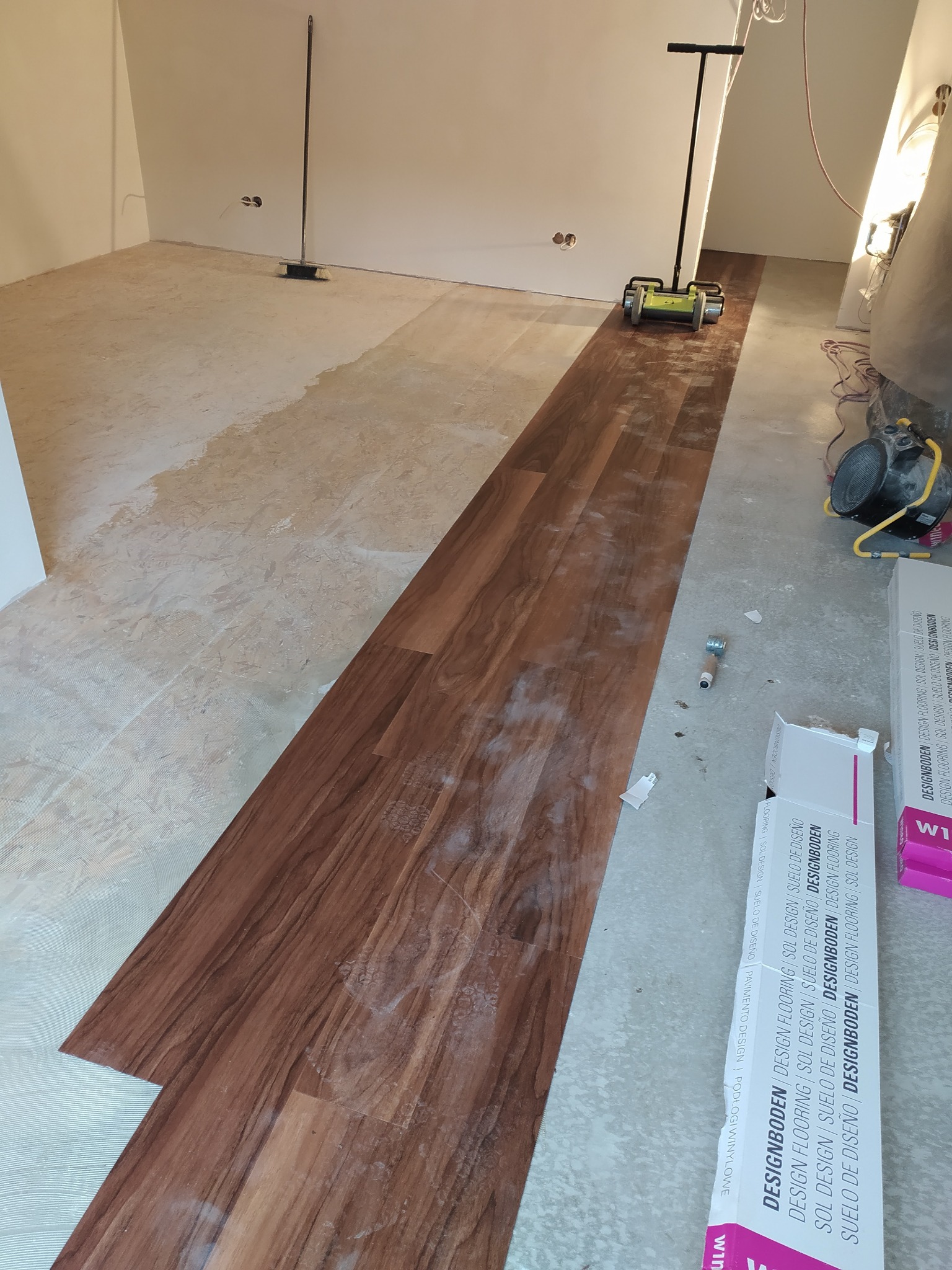 Układanie paneli podłogowych w pomieszczeniu. Częściowo ułożone panele w odcieniach brązu, widoczny sprzęt do montażu i opakowania paneli. Praca w toku.