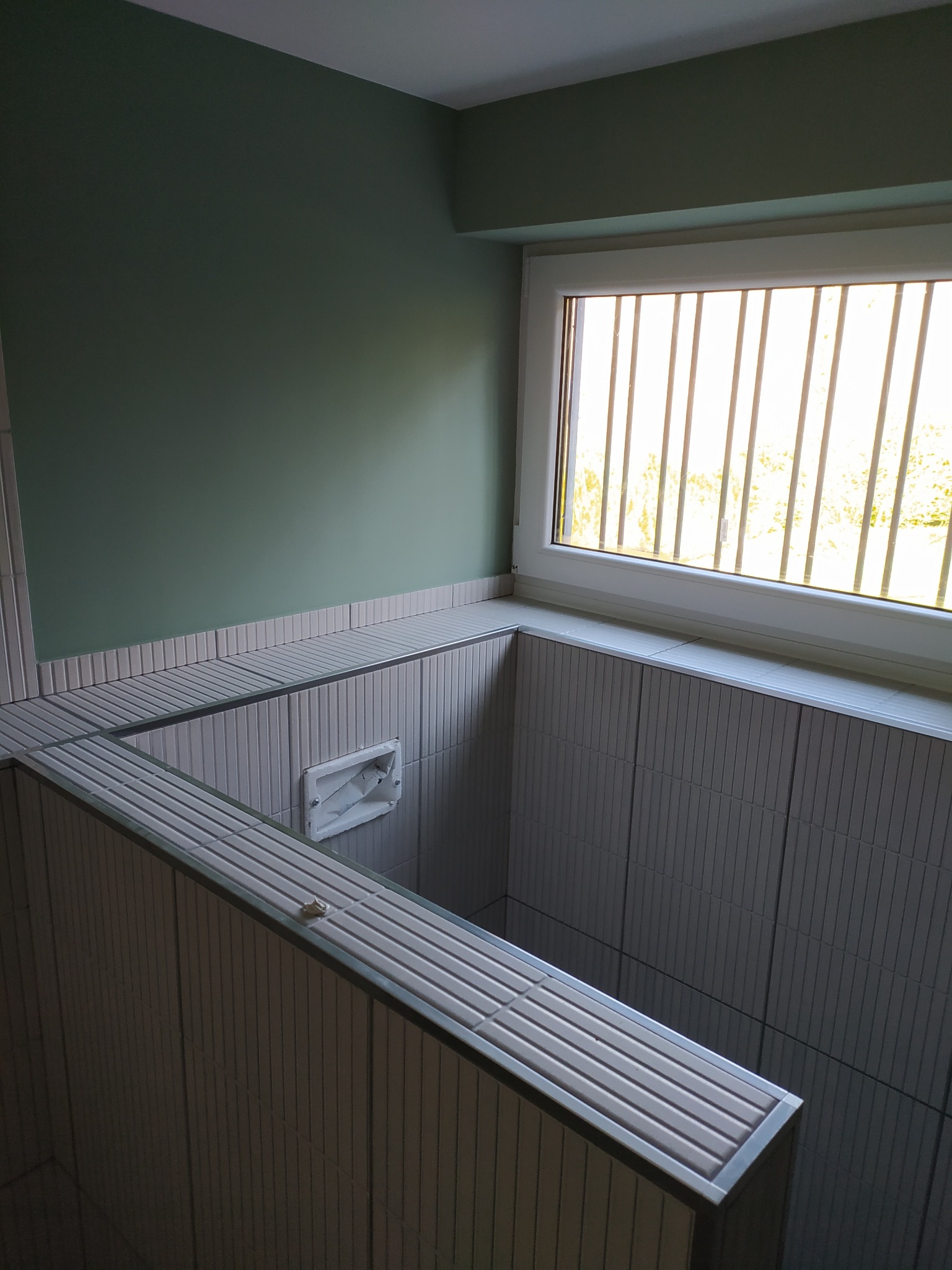 Nowoczesne wykończenie wnętrza z zieloną ścianą i oknem z kratami. Uwagę przykuwają płytki w pionowe paski, tworzące unikalną strukturę. Minimalistyczny design.