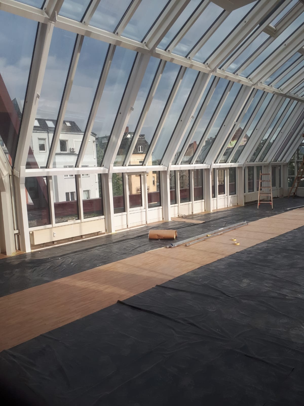 Wnętrze oranżerii w trakcie prac wykończeniowych: podłoga zabezpieczona folią, widoczne elementy konstrukcyjne dachu i okna. Na podłodze leżą narzędzia i materiały.