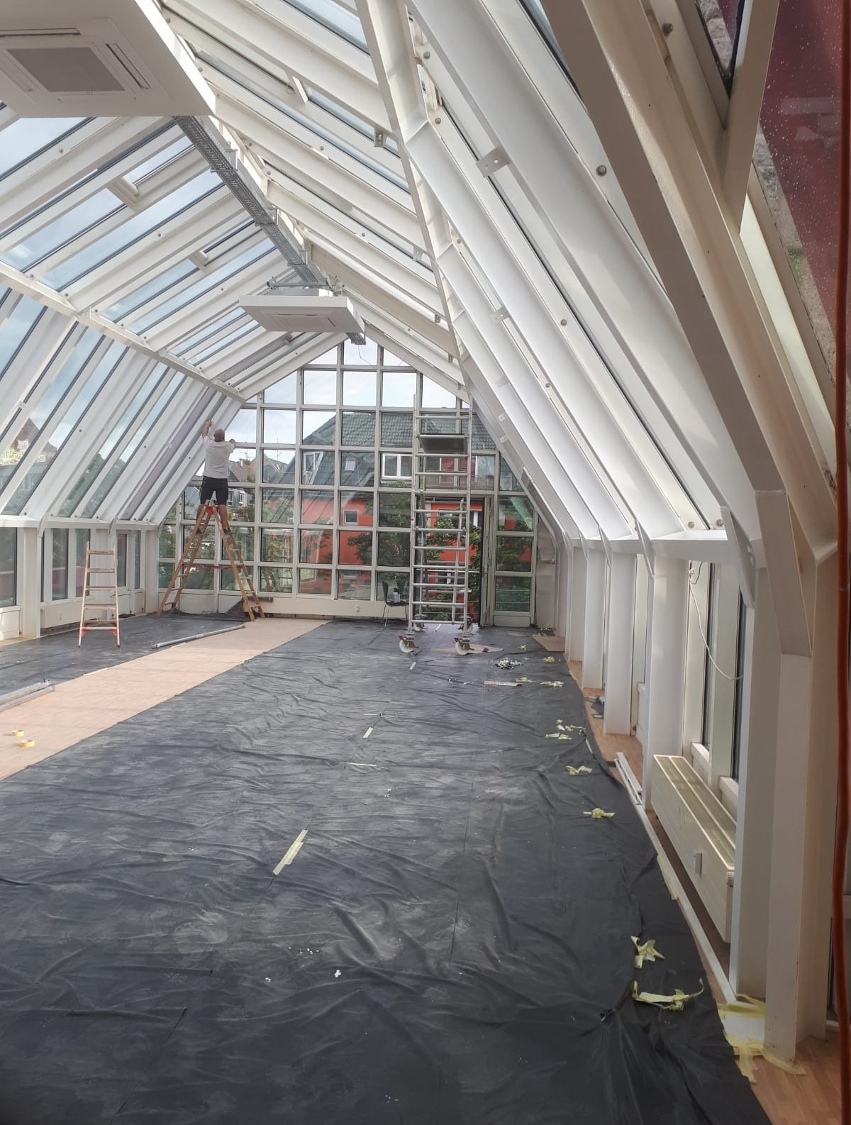 Wnętrze oranżerii w trakcie prac wykończeniowych: malowanie konstrukcji szkieletowej. Podłoga zabezpieczona folią, drabiny w tle, widoczny pracownik na drabinie.