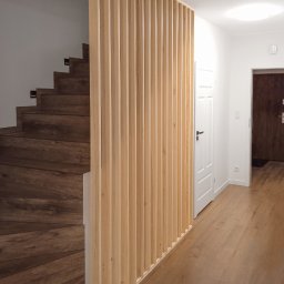 Wojciech Raś Twój Wymiar Pracownia Mebli - Drewniana ścianka działowa z pionowych listew, oddzielająca schody od korytarza. Naturalne drewno, minimalistyczny design, ciepłe oświetlenie.