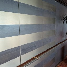 Wojciech Raś Twój Wymiar Pracownia Mebli - Panel ścienny z poziomymi pasami w odcieniach szarości i srebra, z metalową poręczą. Widok z boku, ukazujący strukturę drewna i połysk metalu.