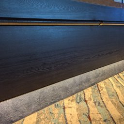 Wojciech Raś Twój Wymiar Pracownia Mebli - Ciemna, drewniana zabudowa z poziomym, metalowym relingiem i subtelnym oświetleniem przypodłogowym, na tle wzorzystej wykładziny. Detal wykończenia wnętrza.