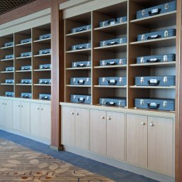 Wojciech Raś Twój Wymiar Pracownia Mebli - Regał z jasnego drewna z szufladami i półkami, na których ułożone są szare walizki. Widoczny fragment wnętrza z dywanem w geometryczne wzory.