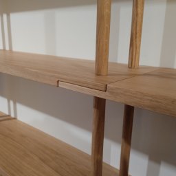 Wojciech Raś Twój Wymiar Pracownia Mebli - Drewniany regał z jasnego drewna dębowego, minimalistyczny design, widoczne detale połączeń elementów, gładka powierzchnia półek, neutralne tło ściany.