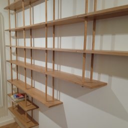 Wojciech Raś Twój Wymiar Pracownia Mebli - Drewniany regał na książki o minimalistycznym designie, z jasnego drewna, zamontowany na białej ścianie. Na półkach leżą książki. Widoczny grzejnik.