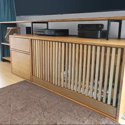 Wojciech Raś Twój Wymiar Pracownia Mebli - Drewniana szafka RTV z lamelami i półkami, na której stoi sprzęt elektroniczny. Widoczny fragment okna z zasłoną i dywan. Unikalny design z otworem na boku.