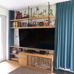 Wojciech Raś Twój Wymiar Pracownia Mebli - Drewniana szafka RTV z lamelami i półkami pod sufitem, na których stoją modele budowli z klocków. Całość w jasnym, nowoczesnym wnętrzu z błękitnymi zasłonami.